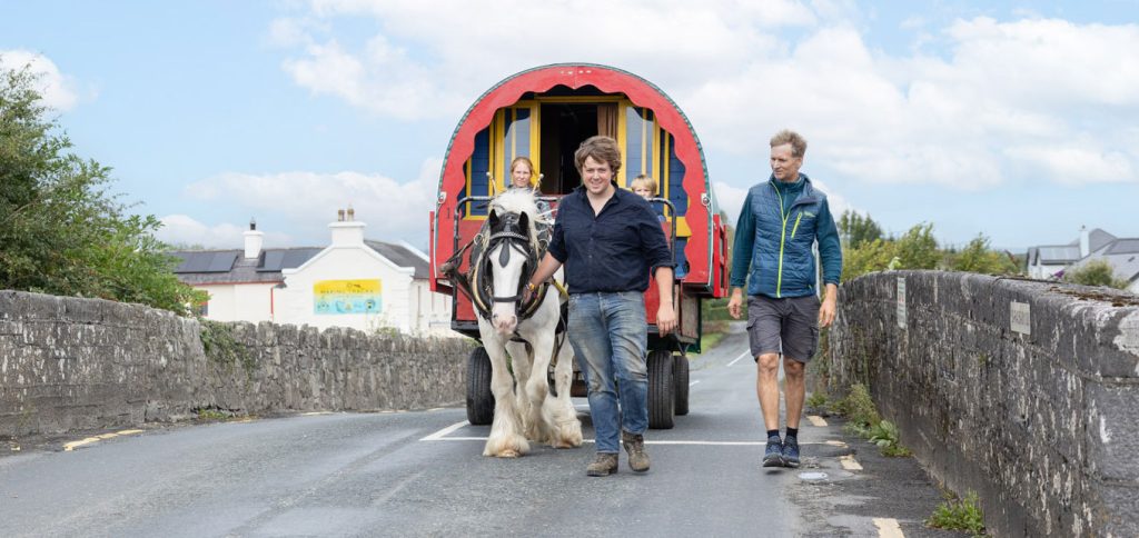 Three people walk with a horse pulling a colorful wagon on a stone bridge.