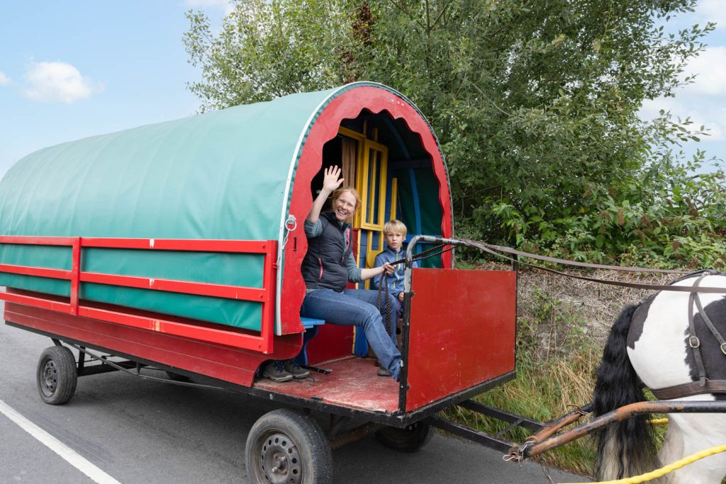 Woman and child wave while sitting in a colorful covered wagon pulled by a horse on a road.