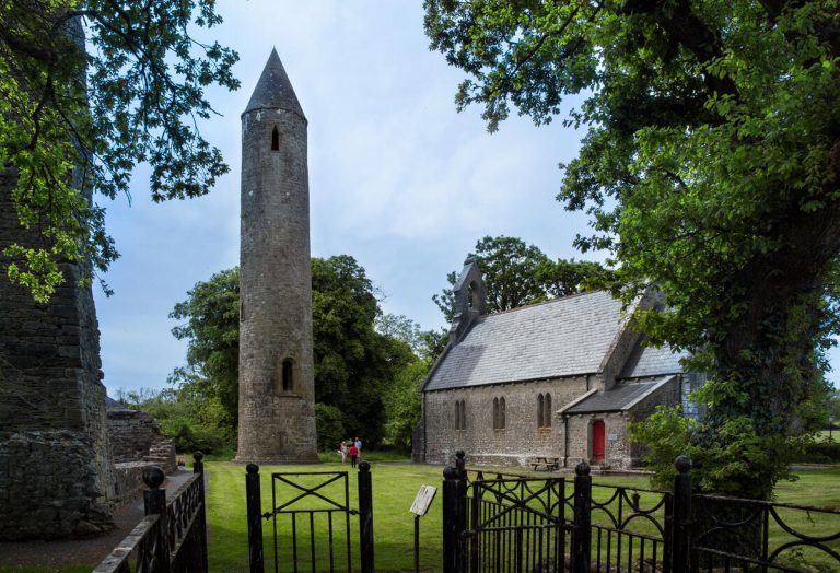 Round stone tower and old church surrounded by trees and a black metal fence on a grassy lawn.