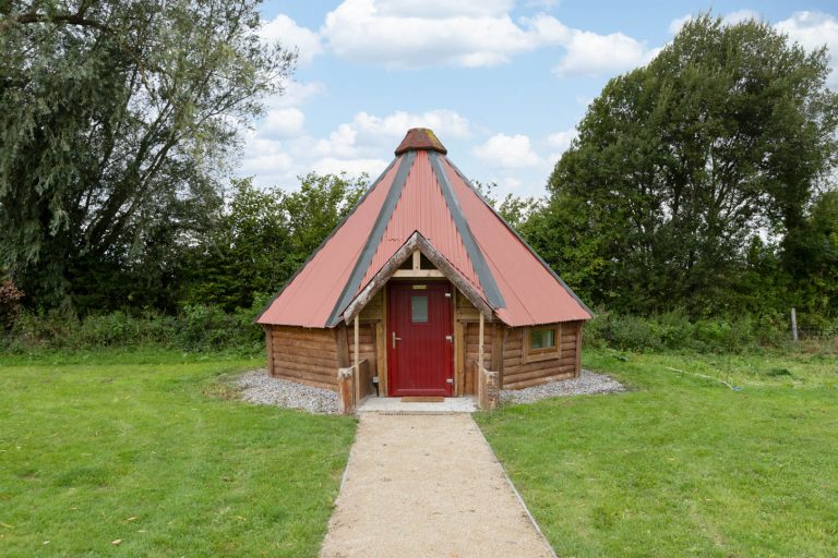 Small wooden cabin with a red roof and door, surrounded by grass and trees.