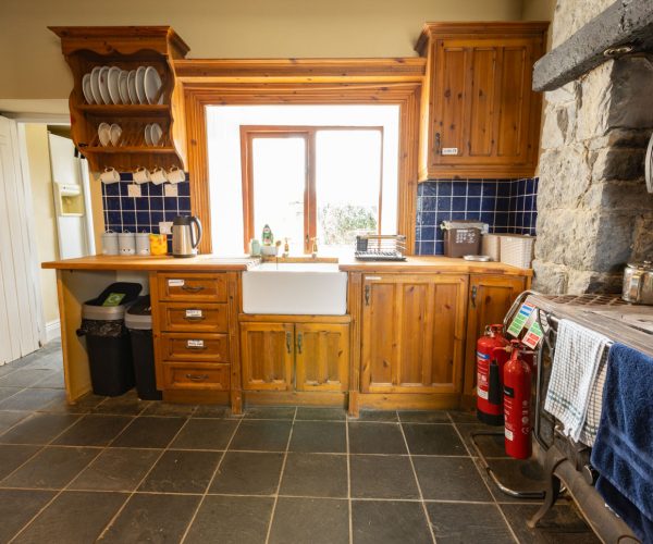 Rustic kitchen with wooden cabinets, sink, dishes, fire extinguishers, and stone wall.