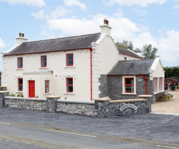 White two-story country house with red trim and stone wall beside a rural road.