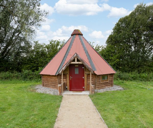 A small wooden cabin with a red door and cone-shaped roof, set on grass with trees behind.
