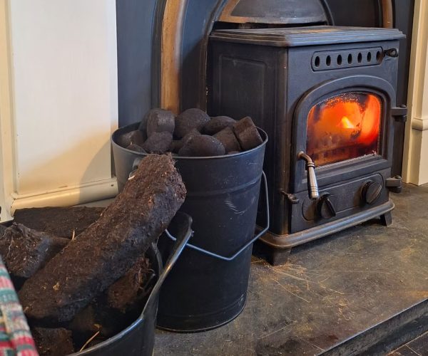 A lit wood-burning stove beside buckets of peat and coal in a cozy room.
