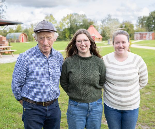 Three people standing outside on grass, smiling at the camera, with buildings in the background.